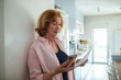 © Marko Geber - Senior woman checking bill while using smartphone in kitchen at home