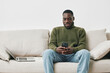 © SHOTPRIME STUDIO - Young African man in a cozy green sweater happily using his smartphone while sitting on a light sofa, with a laptop beside him, reflecting modern lifestyle