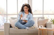 © Pixel-Shot - Happy young African-American woman with basket of ice cream sitting on sofa in living room