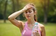 © miss irine - Young woman experiences heatstroke in sunlight. She holds bottle of water in hand. Overheated female covering head, feeling bad. Summer heat, sun, thirst, exercise, outdoor sport.