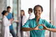 © fizkes - I love my work. Portrait happy smiling enthusiastic female doctor nurse in blue surgical scrubs look at camera form heart shape with fingers mean passion for career job ready to help support patients