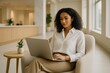 © Darya - A woman with curly hair sits comfortably in a stylish armchair focused on her laptop. Natural light fills a contemporary office space, highlighting her concentration