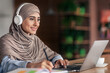 © Prostock-studio - Smiling muslim lady studying online at cafe, using laptop and headset, free space. Joyful female student having online lesson, looking at laptop screen and taking notes, online education concept