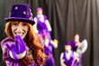© JoseIMartin - Redhead woman wearing purple top hat and gloves smiles and gestures invitingly with other circus performers standing behind her out of focus