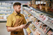 © Seventyfour - Bearded store employee wearing apron checking inventory on digital device while standing in front of products in grocery store aisle during work shift
