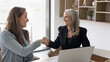 © fizkes - Two smiling businesswomen of diverse generations handshaking at office desk greeting with formal introduction gesture express mutual respect trust feel satisfied with deal teamwork reaching agreement