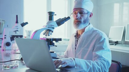 Wall Mural - mature male scientist in white coat using microscope and typing on laptop, conducting research in mo