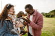 © Drobot Dean - A woman in her late 20s in a denim jacket holding a dachshund in a sweater while standing with a Black male friend in his 20s in a pink jacket petting a dog in a park with a grass field and trees