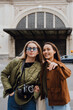 © Drobot Dean - Two smiling White women in their late 20s stand on a street, one White blonde wearing a green jacket and a camera hanging from her neck, and an Asian brunette in a brown jacket who points forward.