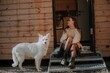 © My Ocean studio - A woman sits on metal steps outside a wooden cabin, enjoying the outdoors with a white dog by her side. She wears a cozy sweater, polka-dot tights, and sunglasses, soaking in the sunlight.