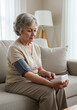 © Gustavo Alves - Senior woman checking her blood pressure at home using a digital monitor. Concept of self-care, preventive health, and independent aging.
