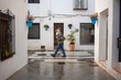 © sutulastock - Elderly man with white beard walking through a charming Spanish street on a rainy day, wearing a colorful sweater, backpack, and cap, surrounded by white buildings and flowers