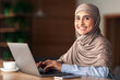© Prostock-studio - Cheerful muslim lady in hijab working online at cafe, using laptop and drinking coffee or tea, copy space. Side view of smiling arab woman freelancer typing on laptop, enjoying her distant job
