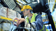 © wkproduction - A man in a yellow hard hat and safety vest operates a forklift in a fast-paced warehouse, highlighting the precision and speed of global supply logistics.