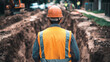 © Oleksandr - A construction worker in an orange hard hat and safety vest surveys a deep trench at a job site, ensuring safety and oversight