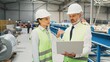 © VAKSMANV - Male and female production engineers having discussion. Two manufactory workers communicating at workshop. Industry workers wearing protective helmets and yellow vests. Metal processing.