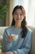 © kineter_ai - Serene Young Japanese Woman in Light Blue Sweater Enjoying a Cup of Tea on a Comfortable Sofa by a Sunny Window: Capturing a Peaceful Moment of Home Relaxation