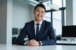 © Alex & M Studio - Poised young East Asian businessman in a suit sitting attentively at a clean white desk in a modern office, offering a confident and professional smile directly to the camera
