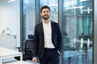 © Liubomir - A smiling businessman stands confidently in a modern office setting, radiating success and professionalism. The natural light enhances the bright, contemporary design.