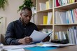 © Dina - diverse male social worker reviewing case files at his desk, focused and dedicated, with office shelves filled with resources behind him, minimal background with copy space