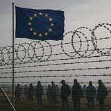 The EU flag waves above a barbed wire fence with silhouettes of people behind it, evoking themes of migration and policy.