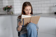© tan4ikk - Sweet Teen Girl Reading Notebook Entries While Sitting On A Chair At Home