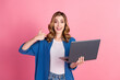 © deagreez - Confident smiling young woman in blue shirt holding a laptop and making a thumbs-up gesture against a pink background.