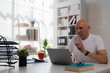 © Tj - Focused mature businessman with finger on lips asking for silence while using laptop computer at workplace, sitting at desk with documents, calculator and coffee cup, bookshelves in background