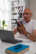 © Tj - Focused mature male freelancer working remotely at a desk in a home office, using a laptop and smartphone while surrounded by books and wearing eyeglasses, showcasing a modern lifestyle