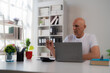 © Tj - Senior manager sitting at desk and working with laptop in bright modern office, he is holding a pen and looking at the screen, plants and books are visible on the shelves