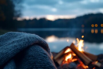 Poster - Cozy outdoor scene featuring a knitted blanket near a campfire with a serene lake and trees in the background creating a calm evening atmosphere