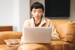 © Iryna - Young woman working on a laptop while sitting on a comfortable couch in a cozy living room during daytime