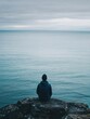 © Aether Pixels - Solitary figure on rocky cliff contemplates vast ocean horizon under overcast sky in serene landscape