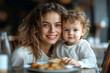 © Infinity Gallery - Young curly haired mother and her little son are having breakfast or lunch in a cafe, sitting at a table. Family weekend