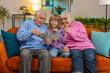 © Andrii Iemelianenko - Caucasian grandfather, grandmother and granddaughter counting money using smartphone calculator at home. Smiling excited girl and old grandparents celebrating success winning lottery sitting on couch