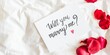 © Rawpixel.com - Romantic proposal card with 'Will you marry me?' text, surrounded by red rose petals on a white bed. Perfect for engagement, proposal, or romantic occasions. Marriage proposal on card with roses.