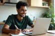 © Vadym - Smiling Indian man uses laptop, studies online, takes notes. Male student studying, e-learning concept. Attractive young man, remote work in home office, education, webinar, conference, work from
