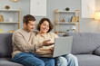 © Studio Romantic - Young smiling couple sitting on sofa in living room at home together watching funny video or using laptop for internet and social media. Happy man and woman resting on couch enjoying weekend