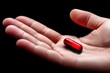 © NickArt - Close-up of a hand holding a shiny red capsule against a black background, symbolizing health and medication.