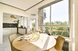 © Casa imágenes - A sunlit dining area featuring a wooden table surrounded by woven chairs, with large windows offering views of greenery, perfect for a cozy dining experience.