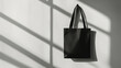 © Bonsales - Monochrome minimalist scene featuring textured tote bag suspended against stark white wall, dramatic light casting sharp window shaped shadow