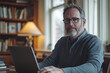 © Nail - A man sitting at a table with a laptop, typing intently. Empty coffee cup and papers scattered around. Bright room with large windows.