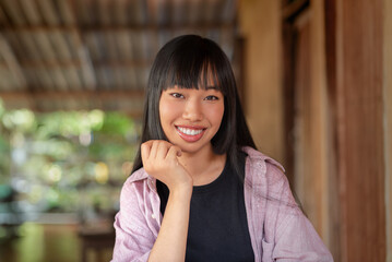 smiling asian woman sitting in rustic wooden home, hand on chin