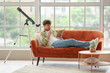 © Pixel-Shot - Young man with telescope and notebook sitting on sofa