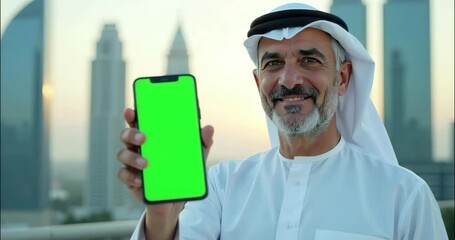 elderly arabian muslim bearded man wearing national clothes, smiling looking at camera and showing empty green screen mobile phone on modern skyscraper city background. mockup for advertising. 4k.