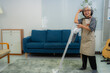 © Tj - Elderly housekeeper wearing headphones and apron using a cordless vacuum cleaner while enjoying music and performing domestic chores in a cozy living room