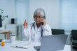 © Tj - Elderly female doctor in a white coat and stethoscope, sitting at her desk, talking on the phone, gesturing while working on her laptop with medical supplies nearby