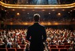 © KarpenArt Studio - a man in a suit stands with his back to the viewer, facing a packed theater with blurred audience members and a glowing stage.