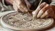 © Husnain - Description: Close-up of experienced hands delicately etching a detailed floral pattern onto a round clay plate, showcasing ceramic artistry.