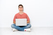 © luismolinero - Teenager Russian man holding pc sitting on the floor isolated on white background and looking up